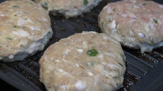 Uncooked chicken burger patties sizzle on black ridged grill, flecked with green herbs and white pieces. Patties slowly cook in place, developing firmer edges over heated grooves as surfaces warm.