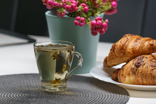 A cup of tea, baked croissants served on a white plate with a warm beverage and a vibrant pink-berried ornamental plant
