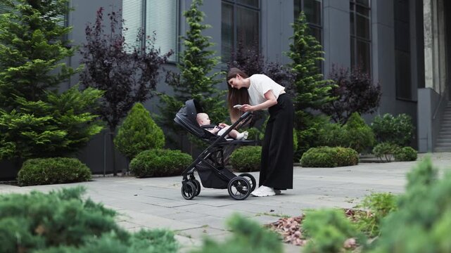 She leans over stroller and reaches toward infant during quiet outdoor moment, with shrubs, trees and paved walkway beside contemporary architecture. Landscaped urban setting in daylight.
