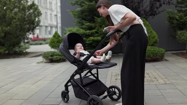 Woman attends to infant in stroller on paved walkway, with close views of baby reclining beneath canopy amid trees and landscaped urban surroundings. Outdoor caregiving moment.