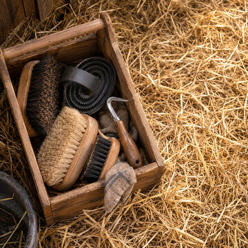 Wooden grooming box with horse brushes, curry comb, hoof pick on golden straw in rustic stable, equestrian equipment