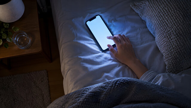 暗いベッドでスマートフォンを見る手元と青い光