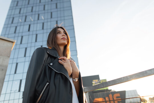 Pensive young woman in a black leather jacket looks up at a modern glass skyscraper from an urban rooftop during golden hour sunset.