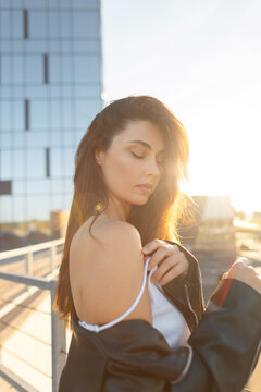Portrait of a young beauty woman with closed eyes, wearing a black leather jacket over a white top and adjusting a strap, against a blurred modern city building at sunset. intimate and serene mood