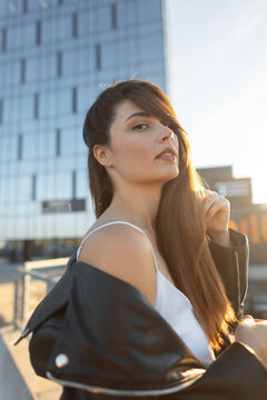 Beautiful young woman with long hair touching her hair, wearing a white top and black leather jacket on a city rooftop during golden hour with a glass skyscraper background.