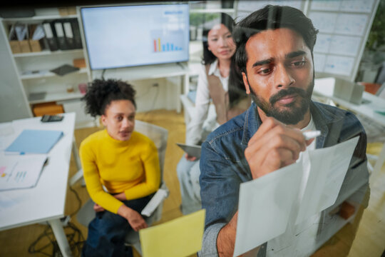 Businessman writing plan on glass board with marker while female colleague watches during office brainstorming