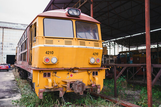 Yellow diesel locomotive parked in railway depot, showing weathered front, metal coupler, headlight, rail track, and industrial maintenance shelter