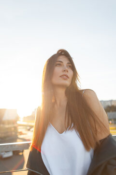 portrait of a beautiful young woman standing outdoors during a golden hour sunset. Backlit by warm sun flare, girl wears a white top and leather jacket, looking off-camera. Atmospheric urban scene