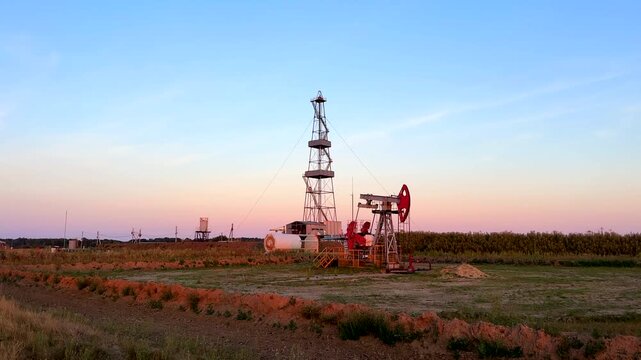 Oil pump jack and drilling rig in open field under sunset sky. Oil extraction from a well. Oil field, oil drilling operations at rig. Crude petroleum industry concept. 