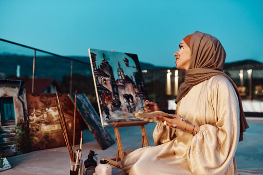 Muslim Woman Artist Painting Cityscape On Rooftop At Dusk Wearing Hijab And Modest Clothing
