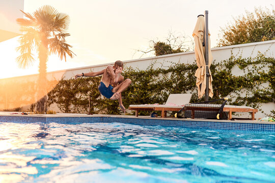 Young Boy Jumping Into Backyard Swimming Pool At Sunset During Summer Vacation