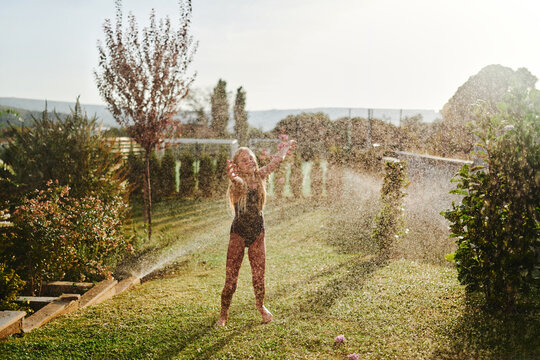 Young Girl Playing In Backyard Sprinkler On Warm Summer Afternoon, Laughing And Enjoying Water