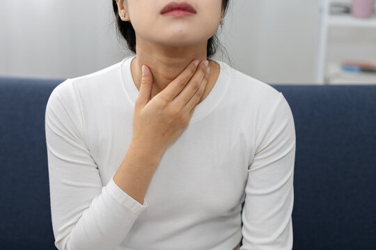 Close up of Asian woman touching painful throat while sitting on sofa at home, suffering from sore throat, neck discomfort, cold symptoms, swallowing pain, respiratory illness.