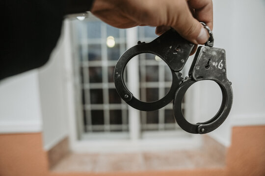 Close-up of a hand holding metal handcuffs with a blurred barred window in the background.