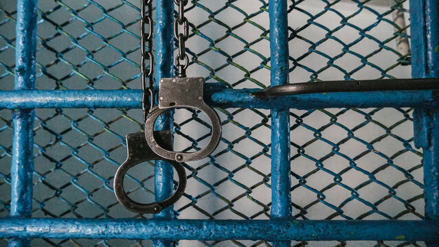 Metal handcuffs hanging on a locked chain-link fence, representing imprisonment, detention and security.