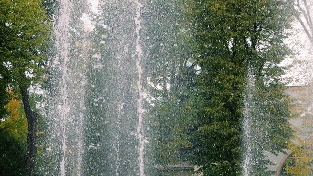 Fountain water jets rise high in slow motion against green trees as crisp splashes create a refreshing liquid wall texture while vertical steady streams spray in the city park.