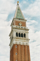 Tall brick bell tower with white marble arches and green patina pyramidal roof topped by gilded statue against cloudy blue sky, closeup vertical view showing ornate cornices, arched belfry openings © Dima Anikin