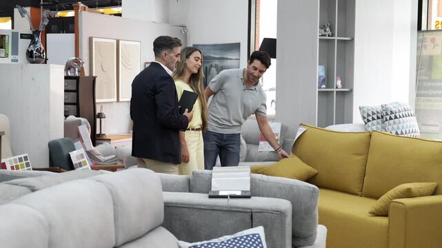 Professional salesman assisting a young couple choosing a comfortable new couch in a modern furniture store. Happy clients buying home decor