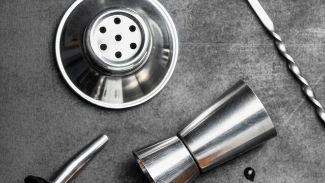 Stainless steel cocktail strainer, jigger and bar spoon on a dark textured surface. Professional bartending equipment for cocktail preparation and mixology