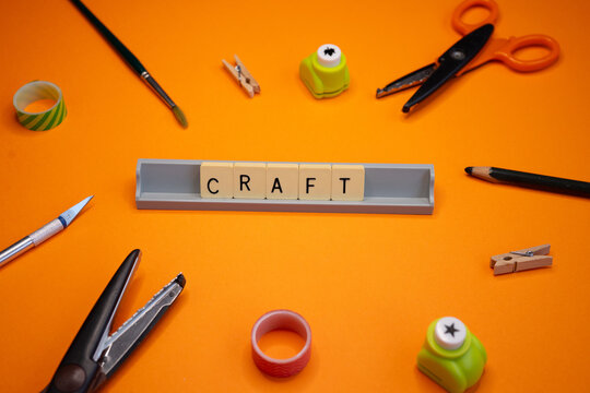 A creative composition viewed from above, featuring craft and stationery tools on a vibrant orange background. Scissors, a craft knife, a paintbrush, a pencil, adhesive tape, and hole punches surround
