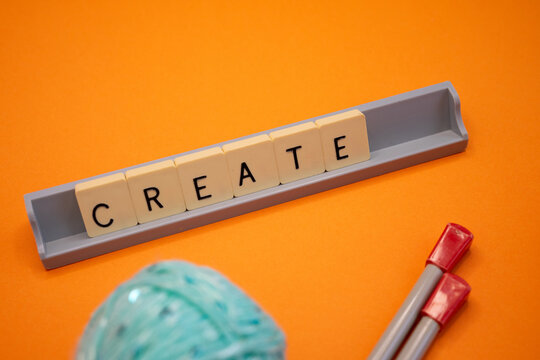 A minimalist, creatively inspired image featuring the word -CREATE- formed by letter tiles, along with a turquoise ball of yarn and knitting needles on a vibrant orange background. Concepts of craftsm