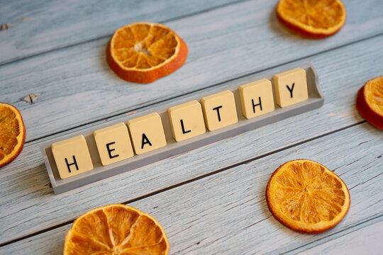 A wide-angle composition on a light-colored wooden surface showing the word -HEALTHY- formed with tiles on a stand. Arranged around the text are several dried orange slices, symbolizing a balanced die