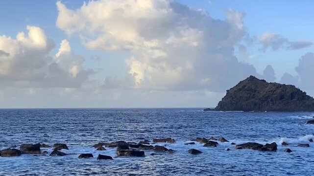 Ocean waves break over dark rocks beside a distant rocky island Roque de Garachico against blue cloudy sky in Tenerife, Canary Islands, Spain,4K.