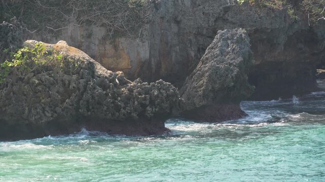 Rocky coastal cliff with large stone formations and ocean waves breaking in tropical turquoise sea water. Scenic wild nature landscape background.