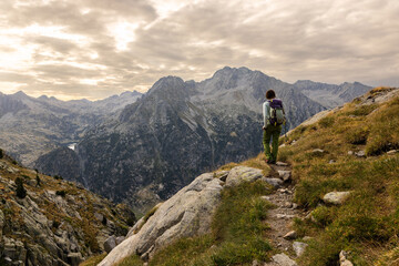 Hiker with a backpack stands on a rocky mountain trail. Misty peaks frame a vast valley under a cloudy sky. They gaze out, calm and contemplative. This scene suits nature, travel, or wellness themes © VICTOR