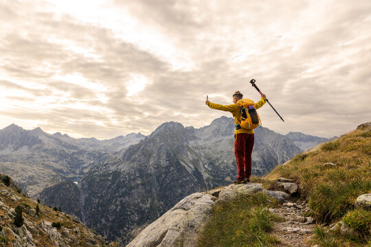 Hiker stands on a rocky mountain ledge. They wear a yellow jacket and red pants. They hold a trekking pole, arms outstretched. Cloudy skies frame distant mountain ranges. Awe and accomplishment