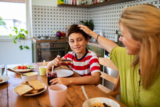 Woman ruffles boy's hair as he eats breakfast