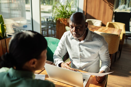 Man explaining document to colleague at laptop in cafe