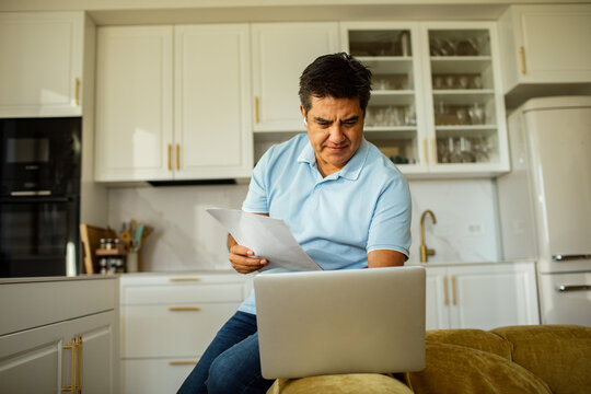 Man reviewing paperwork on laptop in kitchen