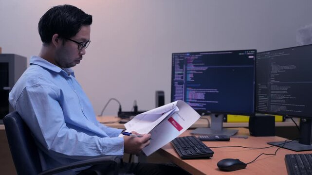 Programmer reads a technical document beside computer monitors displaying source code, representing software development, coding research, debugging, IT workflow, problem solving, and technology.