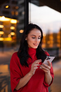 Young woman in red sweater enjoying connecting on smartphone, smiling while scrolling social media in city at evening, urban communication technology concept