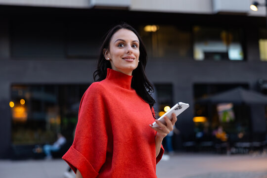 Young woman looking up with confidence while holding a smartphone, standing on an urban street, enjoying city life and modern communication in the evening