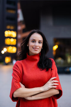 Confident business woman standing with crossed arms, smiling at camera against a modern city building at twilight, symbolizing professional success and ambition