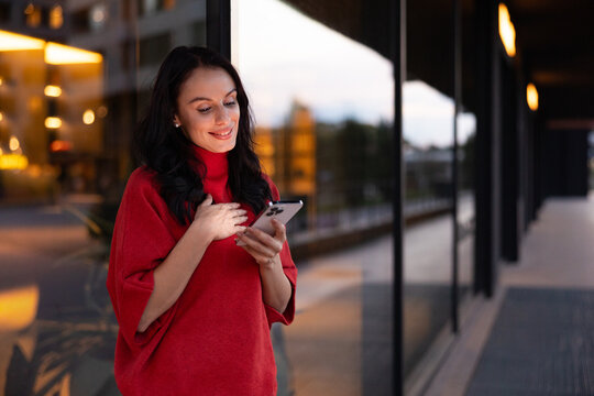 Woman feeling connected and happy while looking at smartphone screen, receiving good news or message, standing in modern urban environment at dusk
