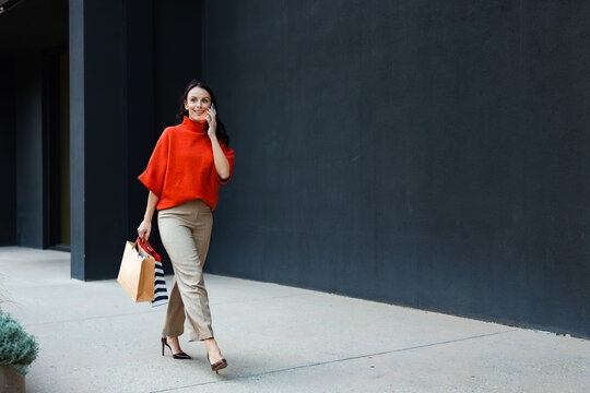 Woman walking outdoors carrying shopping bags and talking on mobile phone, enjoying modern urban lifestyle and retail therapy after successful purchasing