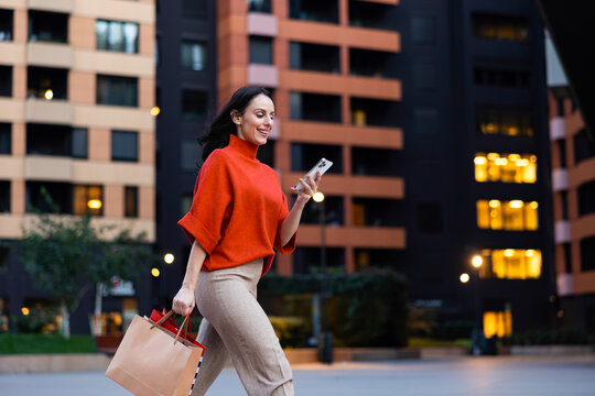 Young woman walking in an urban setting, carrying shopping bags and smiling while using her smartphone, embodying modern consumerism, technology, and city life