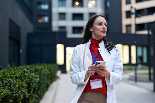 Female doctor wearing a lab coat and stethoscope, holding a smartphone, looking away in a thoughtful moment outdoors, connecting healthcare with technology in an urban setting