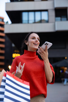 Smiling woman recording a voice message on her smartphone while carrying shopping bags, communicating wirelessly and enjoying a day out in a modern urban environment