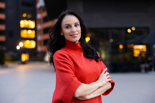 Professional brunette woman wearing a red sweater standing confidently in an urban setting at dusk, expressing elegance, leadership, and determination