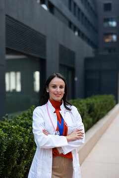 Young female doctor in white lab coat with stethoscope standing confidently outside a modern medical facility, smiling and projecting trust, professionalism and patient-focused care