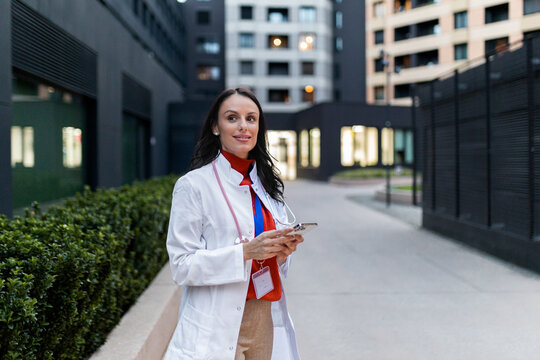 Female doctor in a lab coat and stethoscope walking in an urban setting, using a smartphone, embodying modern healthcare communication, innovation, and professional mobility