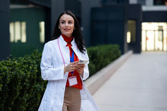 Female medical professional walking outdoors on a modern campus, using a smartphone for communication and digital healthcare applications, embodying innovation in medicine and technology