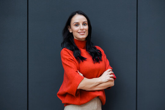 Confident young woman smiling, crossing arms, looking at camera against dark grey wall, portraying professionalism, leadership, and success in business and career