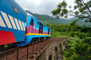 A blue and white passenger train rounds a curved stone viaduct above a forested valley, surrounded by dense tropical trees and green mountains in Vietnam. © aapsky