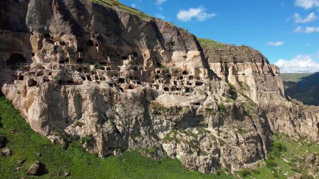 Aerial view moving alongside the ancient cave city of vardzia, a historic monastery complex carved into the erusheti mountain, showcasing its many levels and dwellings under a blue sky