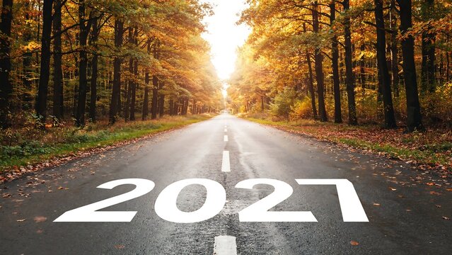 Road with 2027 painted markings extending through warm autumn woods with golden sun glare at horizon for future business goals and new year planning templates.